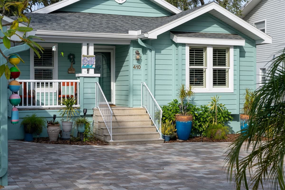 This bright and airy home showcases a stunning paver driveway, charming porch seating, and tropical touches throughout.