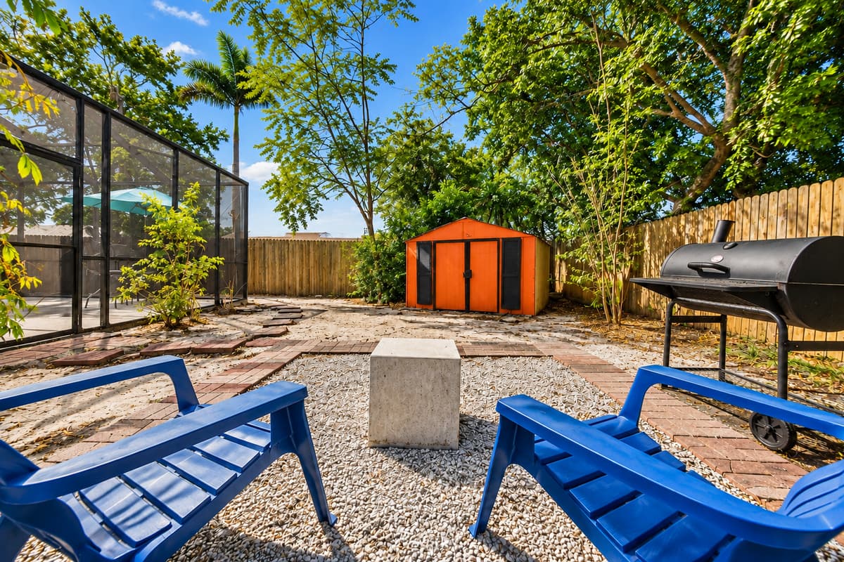 Scenic backyard view showcasing the fire pit seating area, grill station, and surrounding tropical landscaping.
