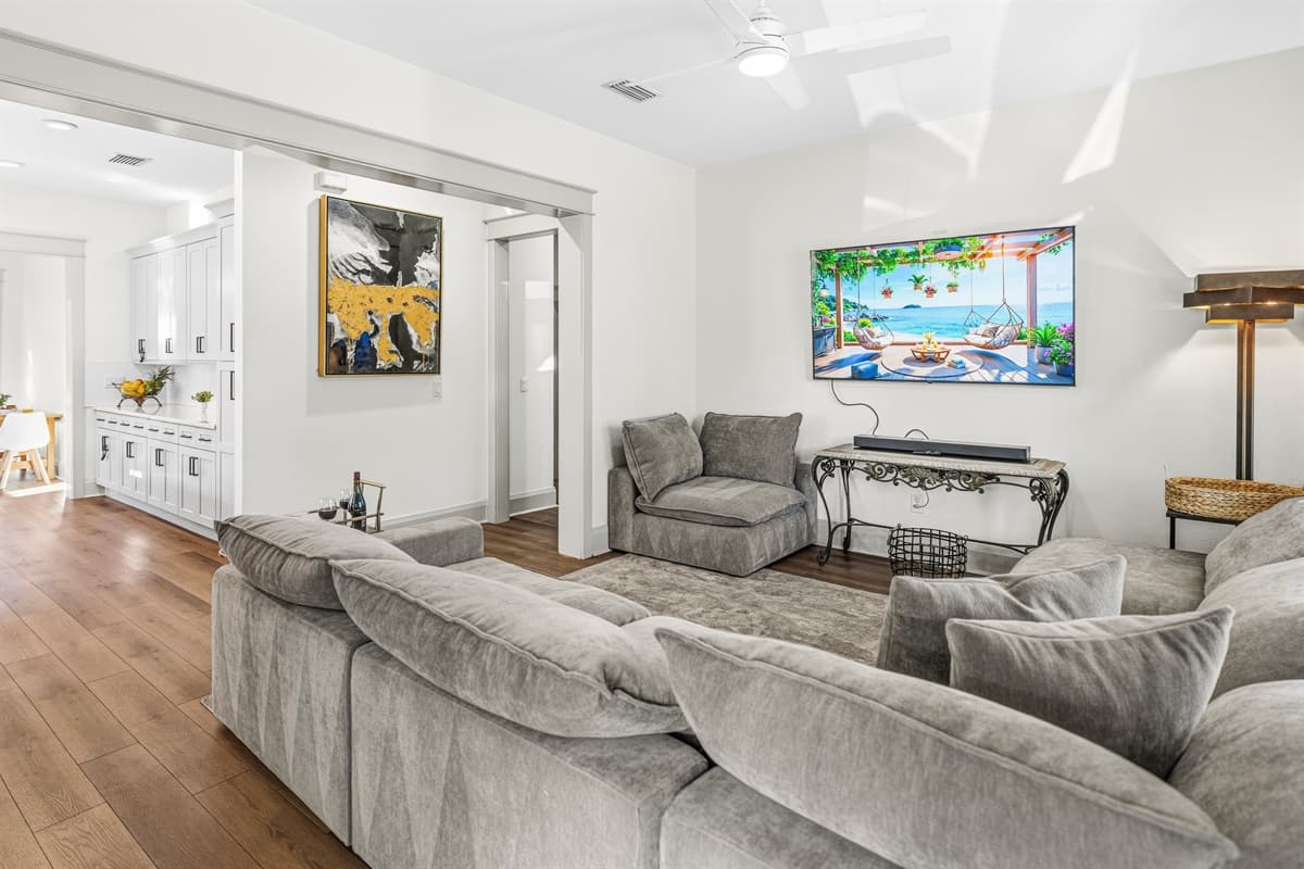 Spacious open-concept living room featuring a plush oversized sectional, gleaming hardwood floors, wall-mounted smart TV, and vibrant tropical artwork. Brand-new construction comfort designed for total relaxation in Seminole Heights.