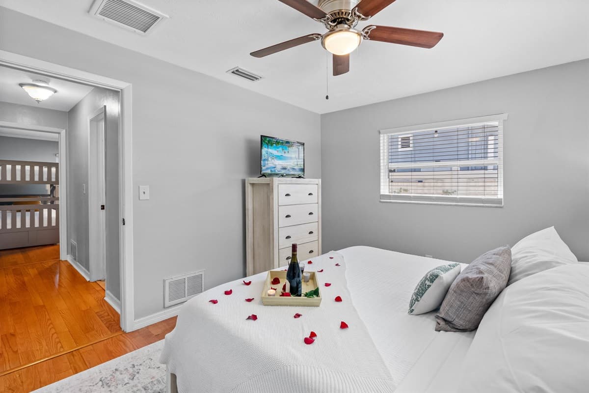 Relax in this peaceful bedroom featuring a crisp white bedspread, soft accent pillows, and subtle beachy decor. A curated tray with wine and candles adds a touch of indulgent charm.