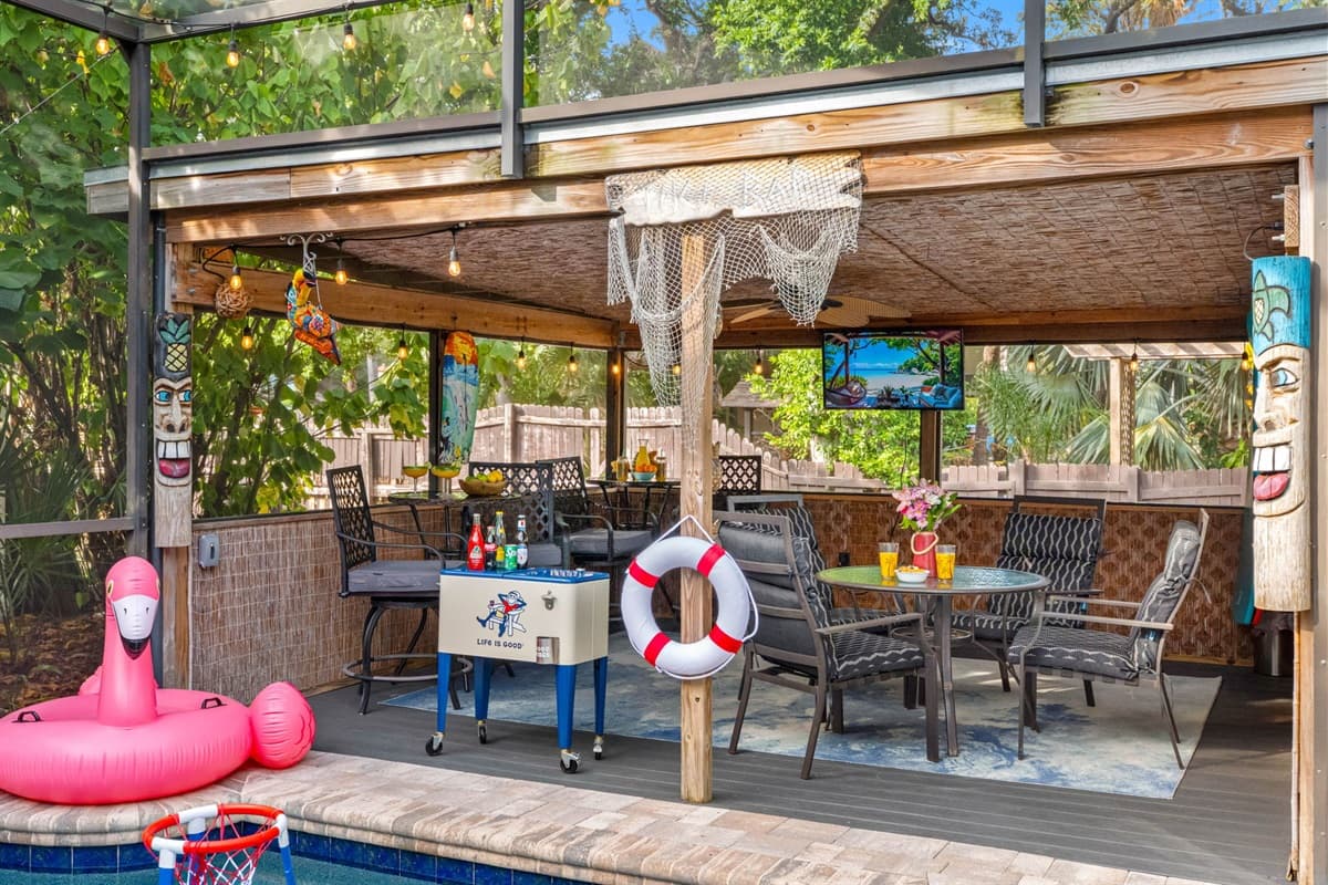 Poolside lounge area with tiki pillars, a beverage cooler, hanging décor, and a shaded dining setup under a bamboo-style ceiling.