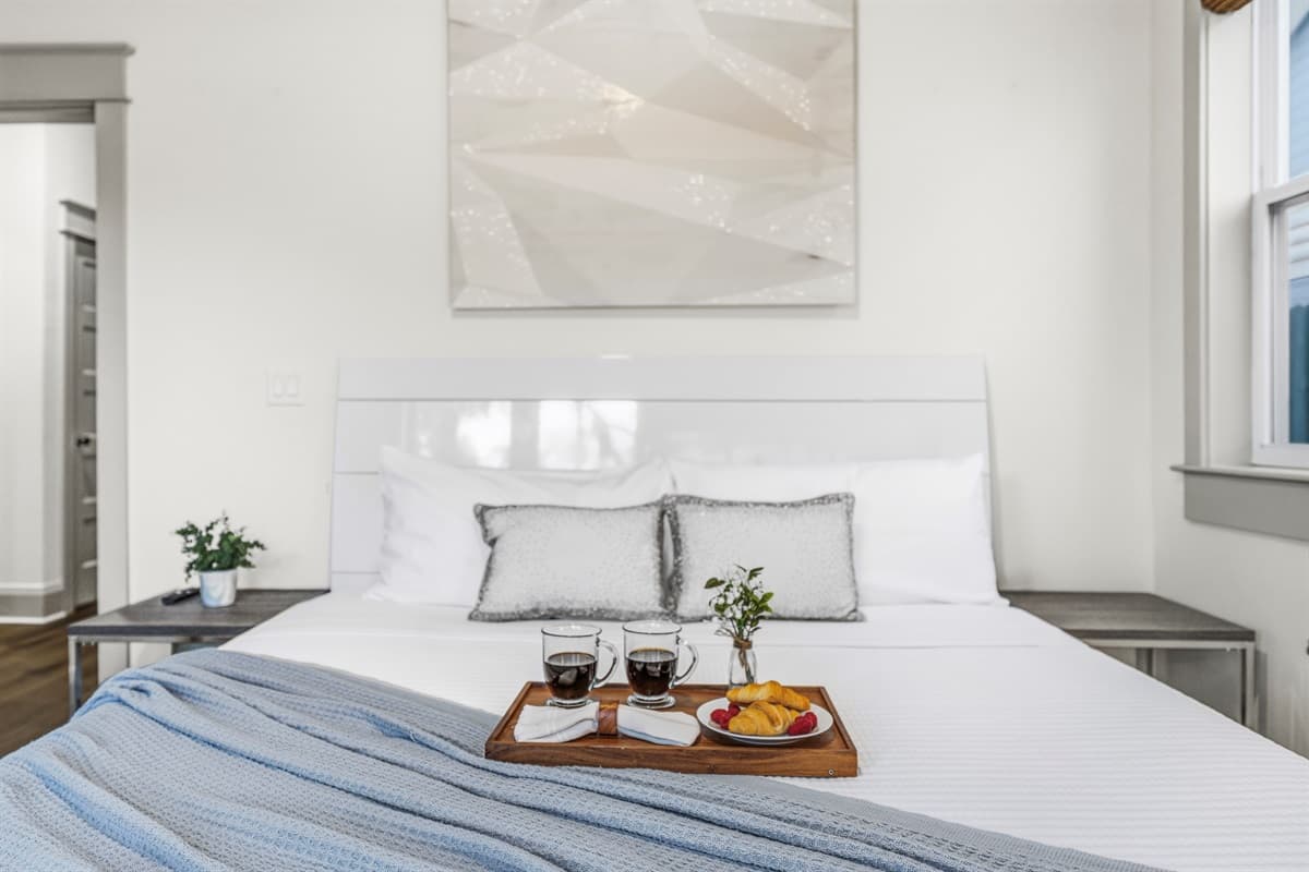 Start your morning right — crisp white bedding, a wooden breakfast tray with coffee and fresh fruit, a potted plant, and soft natural light. Brand-new, hotel-quality comfort in this Seminole Heights Airbnb.