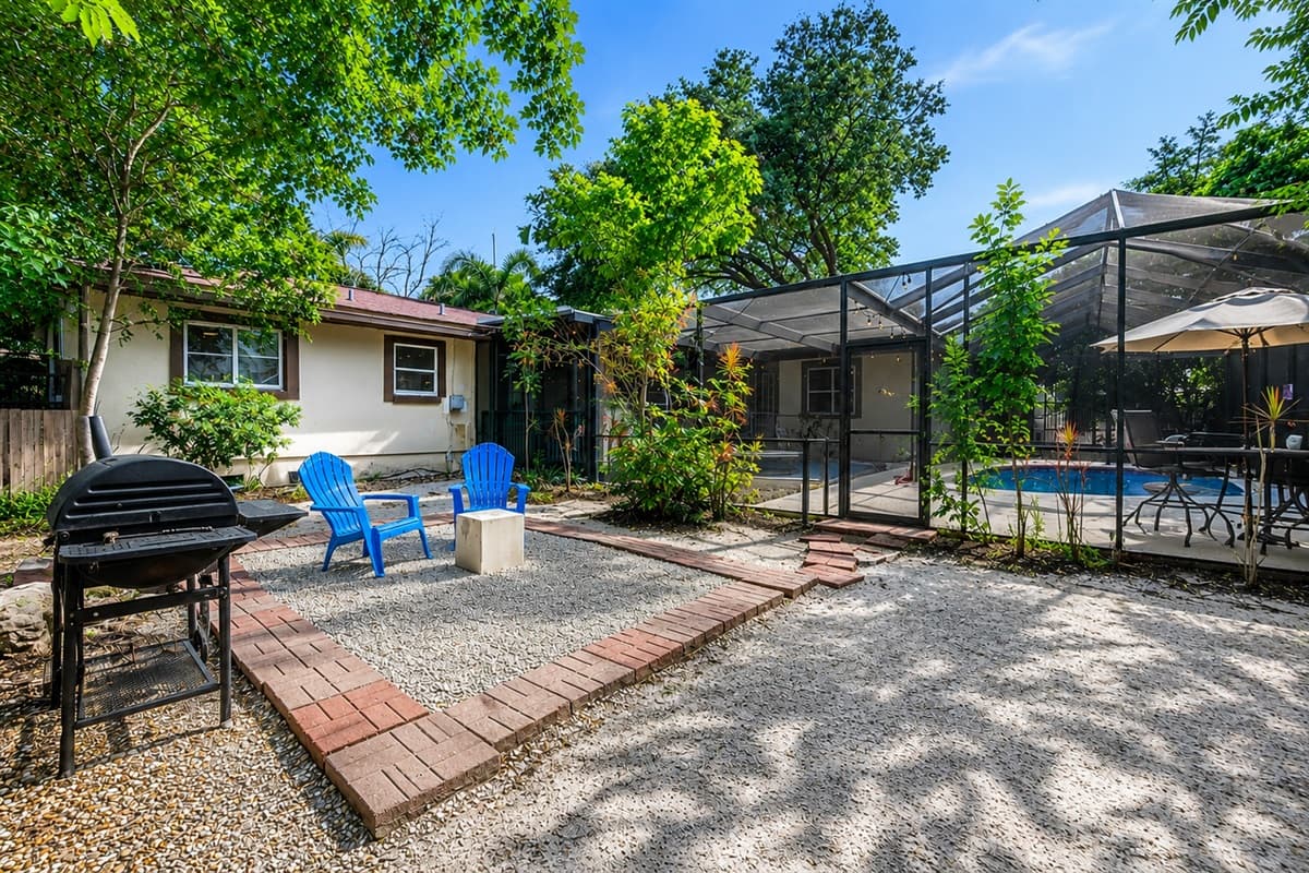 Wide view of the backyard featuring multiple outdoor gathering spaces, mature trees, and screened pool access.