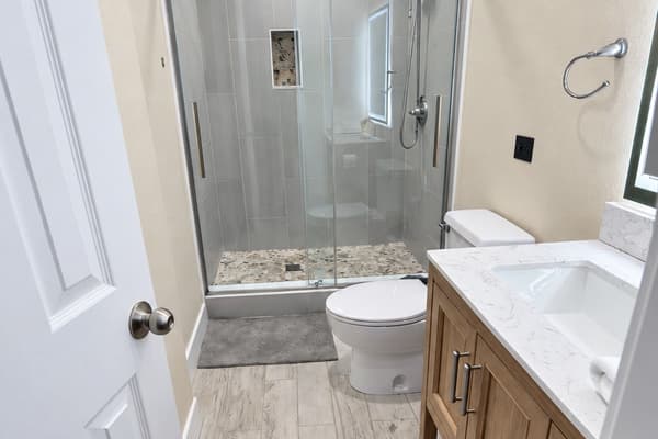 This beautifully updated bathroom features a sleek glass-enclosed shower with pebble tile accents, a modern wood vanity with a quartz countertop, and elegant brushed nickel fixtures.