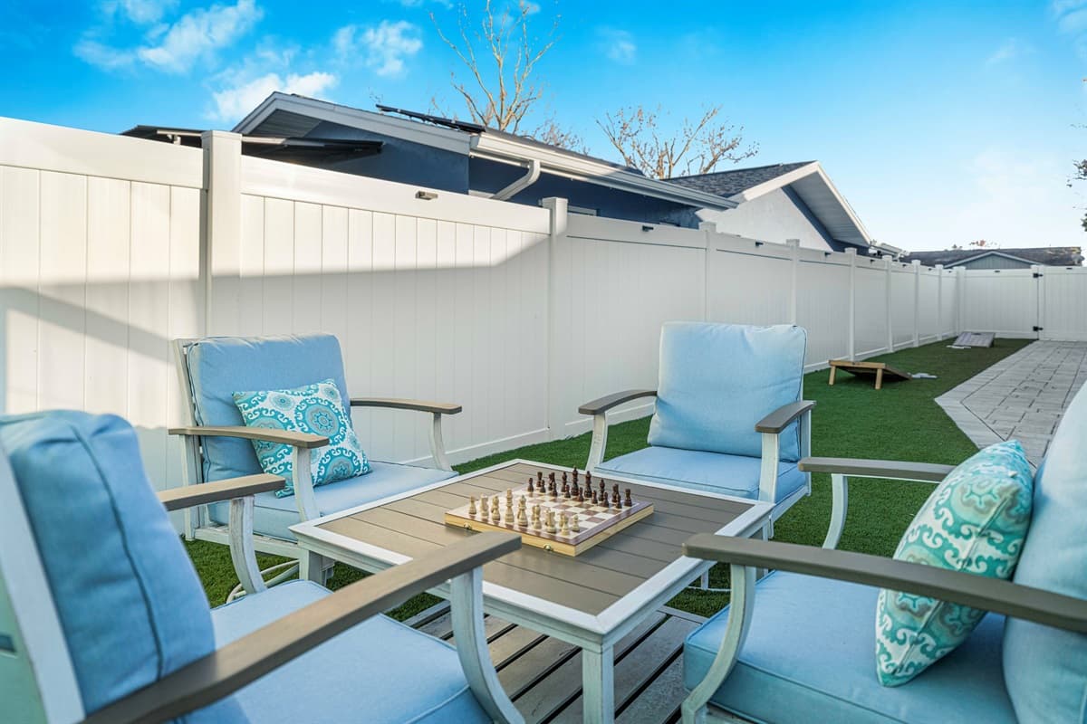 A cozy outdoor seating area with cushioned patio chairs arranged around a small table featuring a chess set. The private fenced yard and neatly kept turf provide a peaceful setting for relaxing or enjoying a friendly game outside.