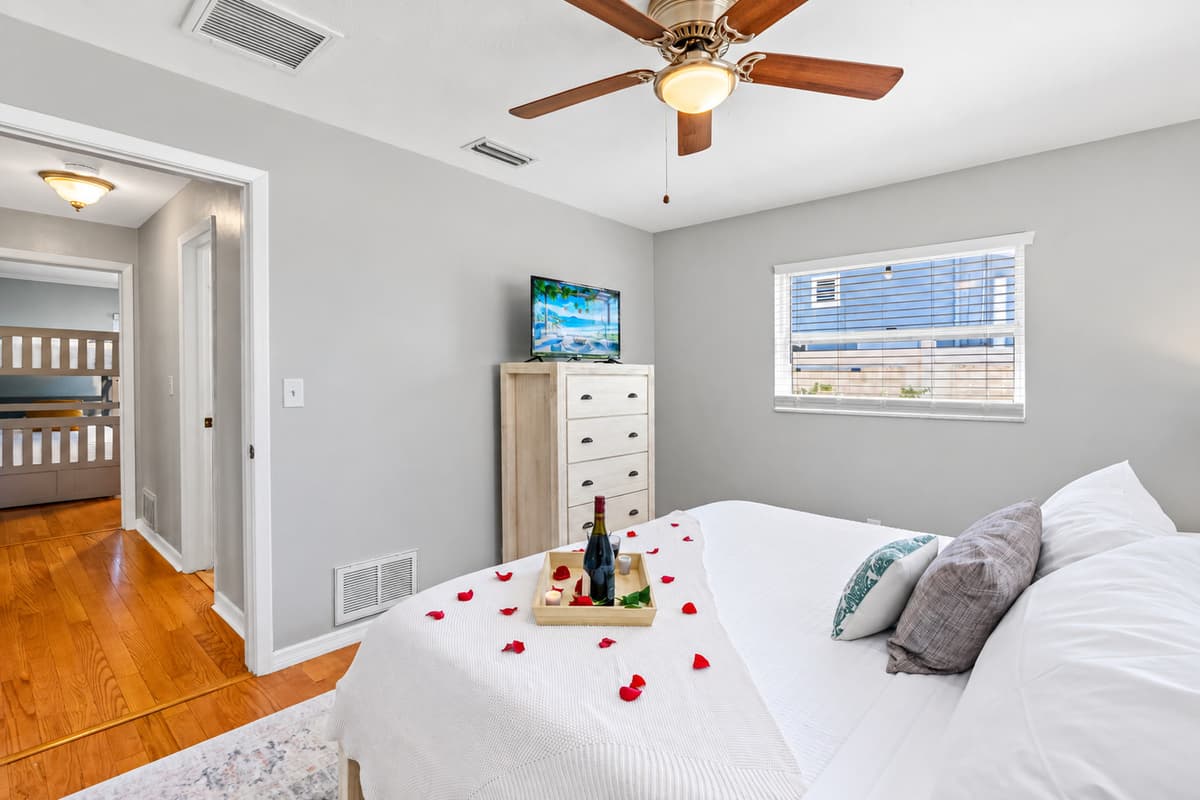 Relax in this peaceful bedroom featuring a crisp white bedspread, soft accent pillows, and subtle beachy decor. A curated tray with wine and candles adds a touch of indulgent charm.