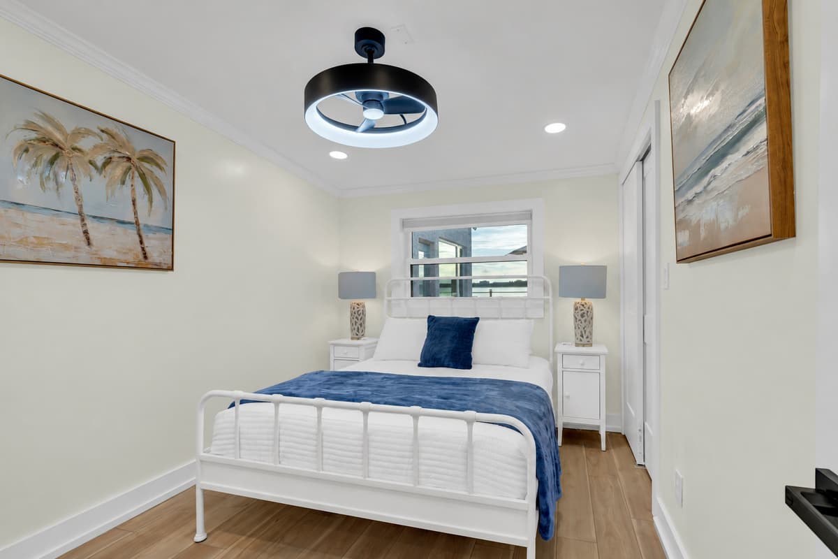 Crisp linens in a breezy queen room. A queen bed with a white metal frame, soft blue bedding and beach art. Simple, fresh and calm, perfect for a couple to recharge.