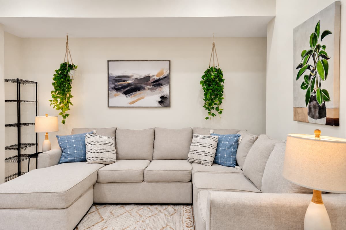 Claim this corner and don't get up until dinner. Oversized sectional with a chaise, warm lamps and green plants — the quiet lounge for reading, napping or waiting out the hottest hour.
