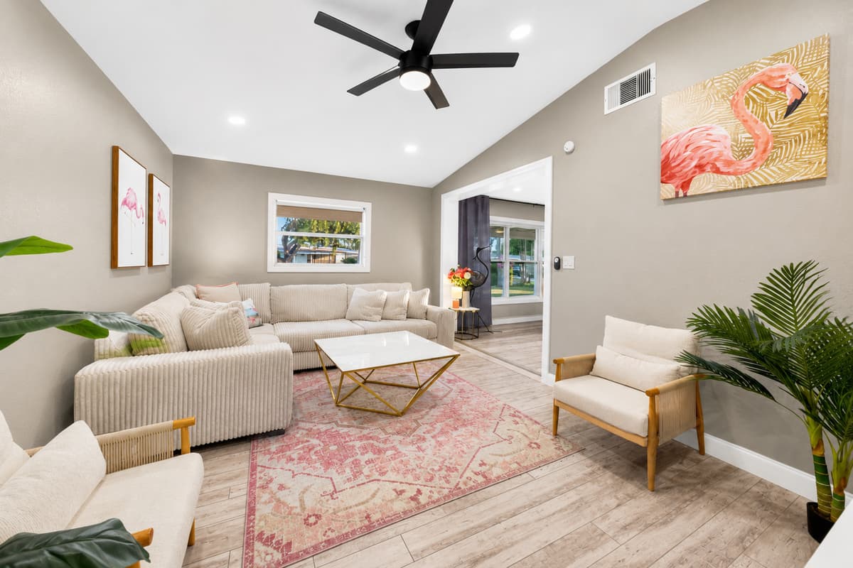 Spacious open-concept living room with neutral furnishings, pink accent rug, ceiling fan, and tropical artwork.